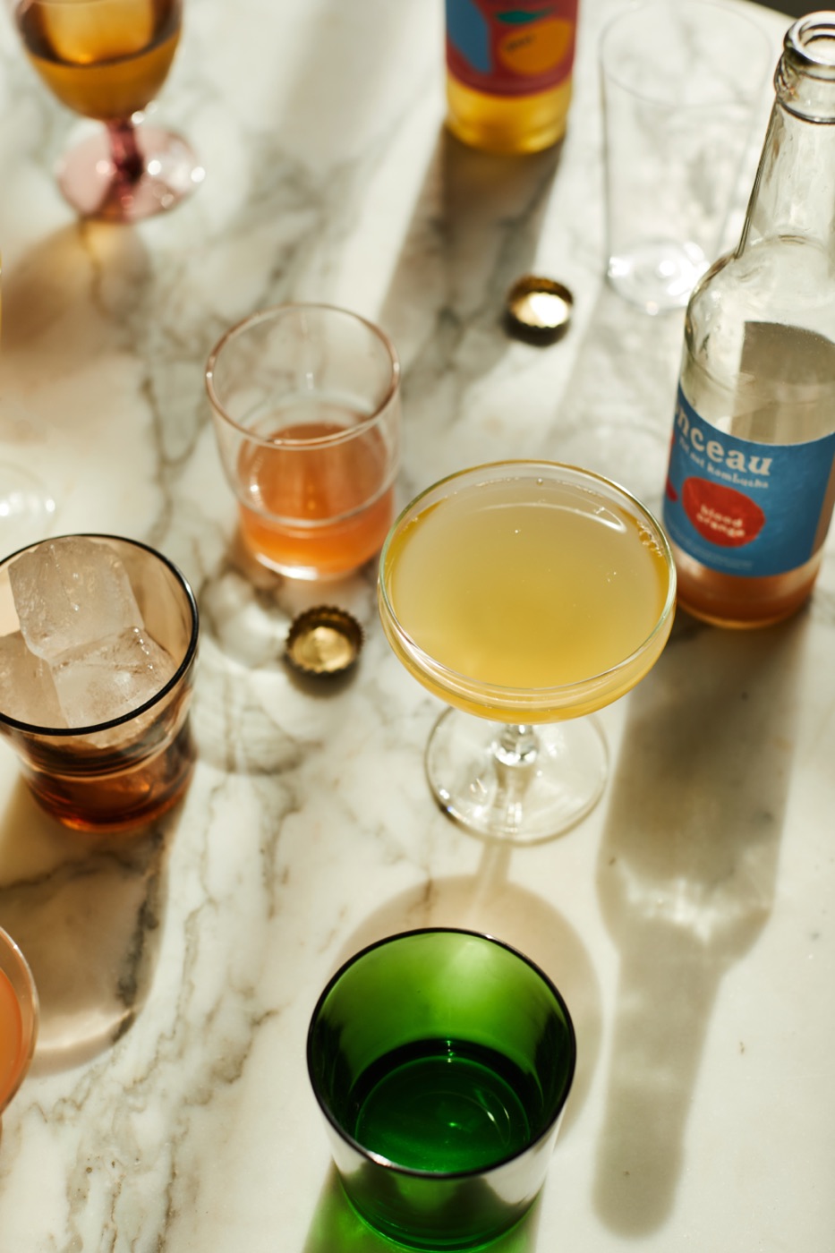 A coupe of pale kombucha, a green tumbler and a Monceau bottle on marble