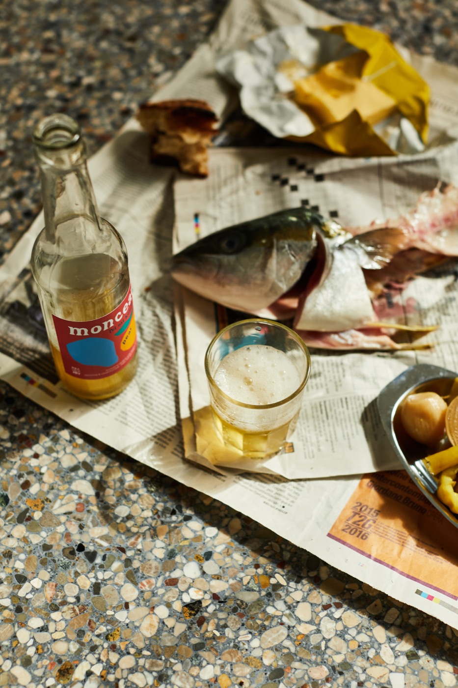 A whole fish on newspaper with a glass of kombucha and a Monceau bottle
