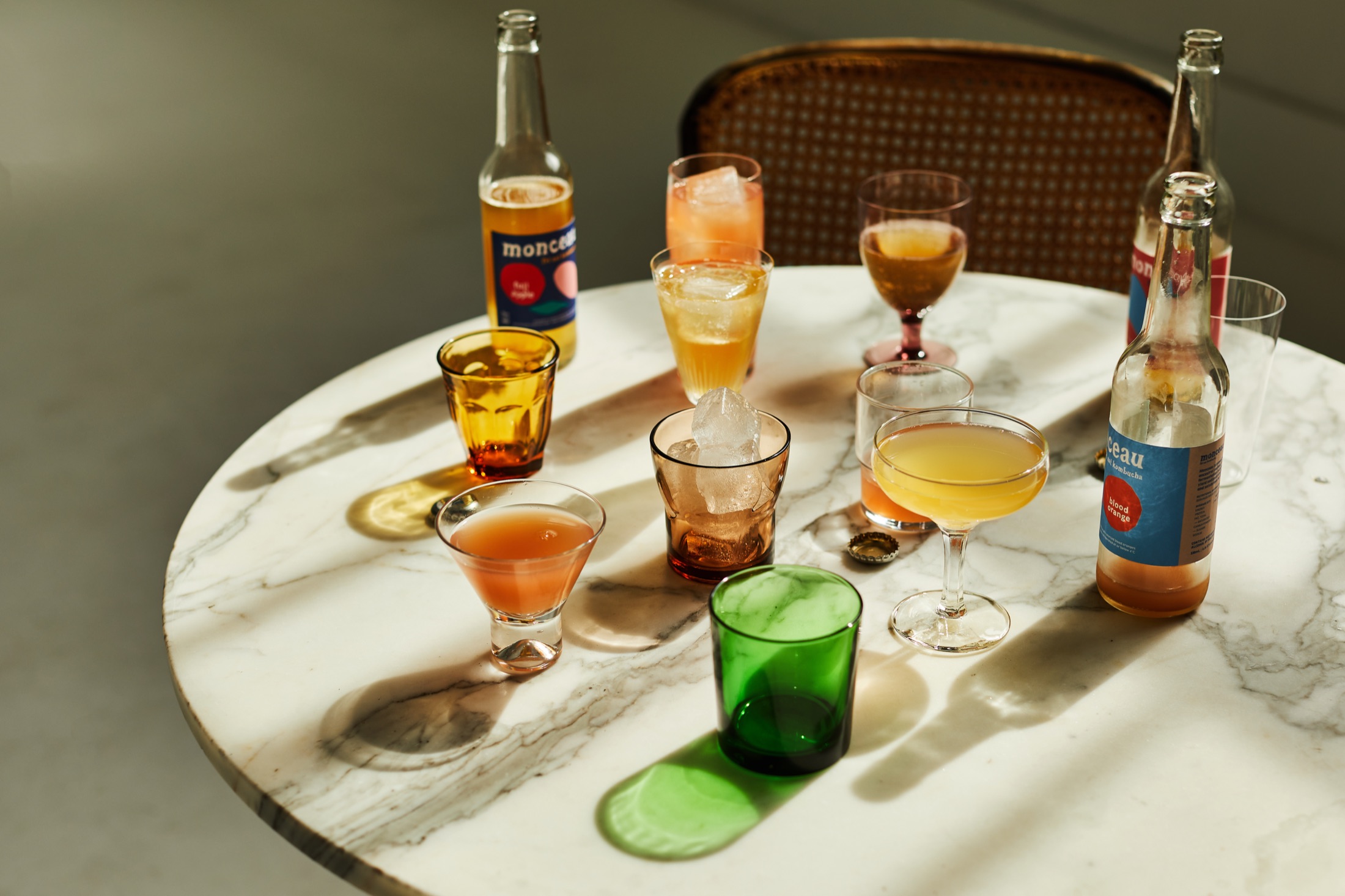 A marble café table in hard afternoon sun, crowded with coloured glasses of kombucha and two Monceau bottles
