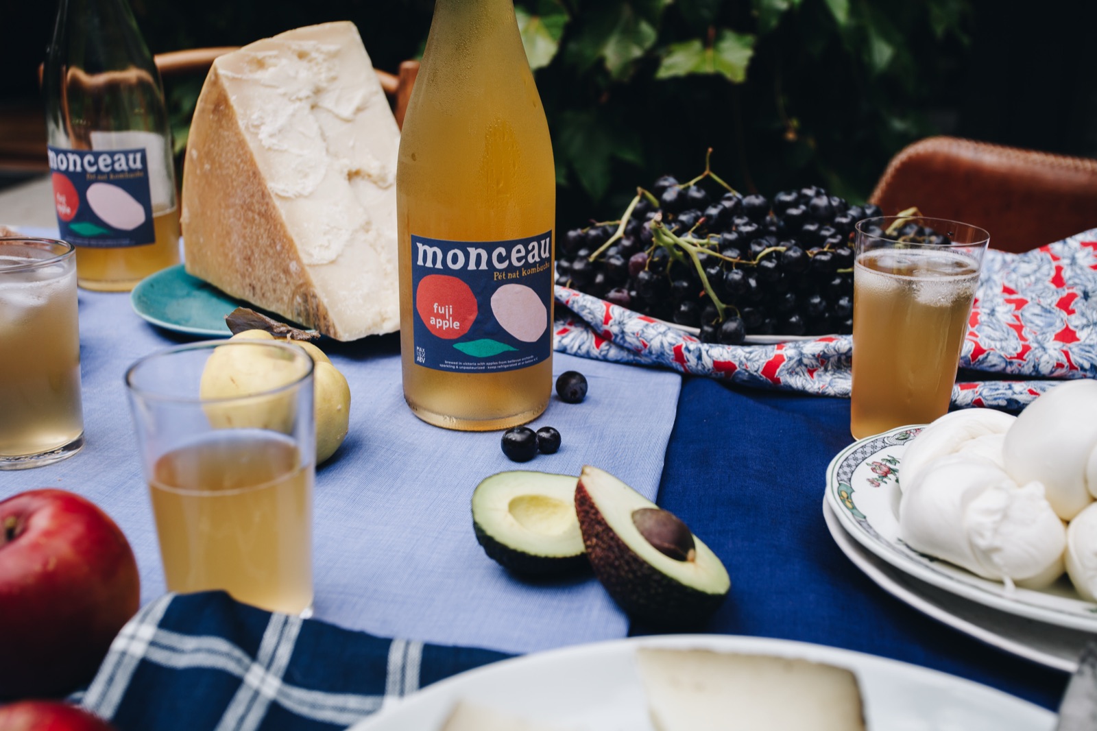 A lunch table with a bottle of Monceau Fuji Apple, cheese, grapes, avocado and blue linen
