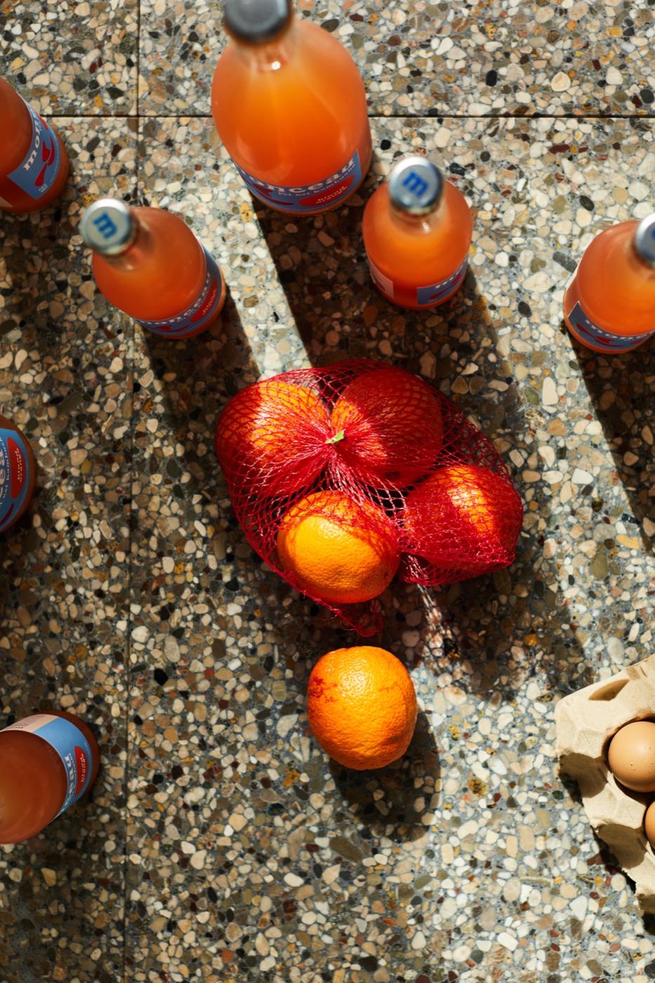 Blood oranges in a red net bag among Monceau bottles on terrazzo
