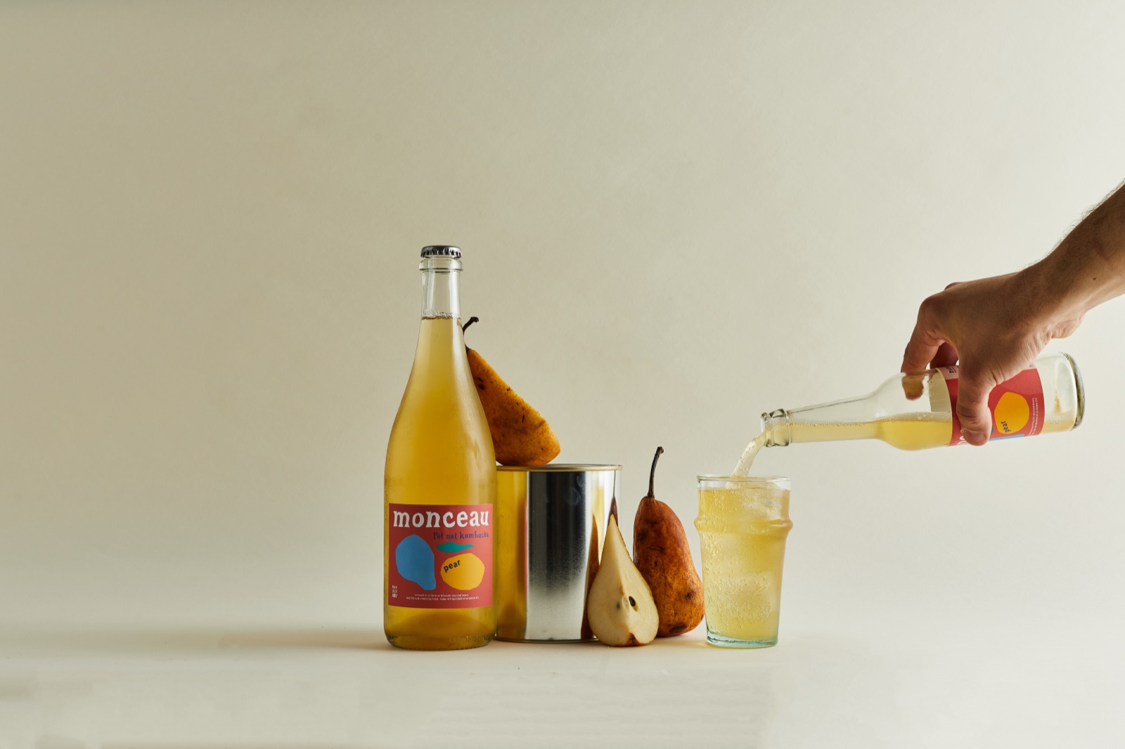 A hand pours pear kombucha into a glass beside a bottle and a ripe pear