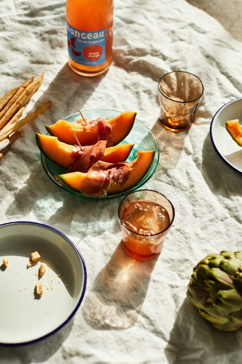 A picnic on a white linen cloth with rockmelon, prosciutto and a bottle of Monceau